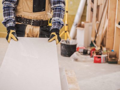 Male Construction Worker Holding Long Piece Of Gray Drywall Material Ready To Be Installed On House Wall.